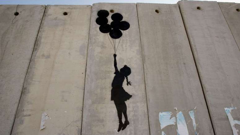 Trabalho de Banksy na parede que separa Israel mostra a silhueta de uma garota suspensa por com bal&otilde;es