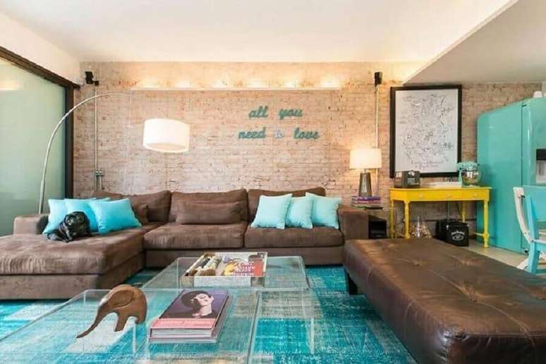 36. Sala de apartamento decorado com parede de tijolinho a vista, tapete azul e sofá marrom amplo – Foto: DT Estúdio