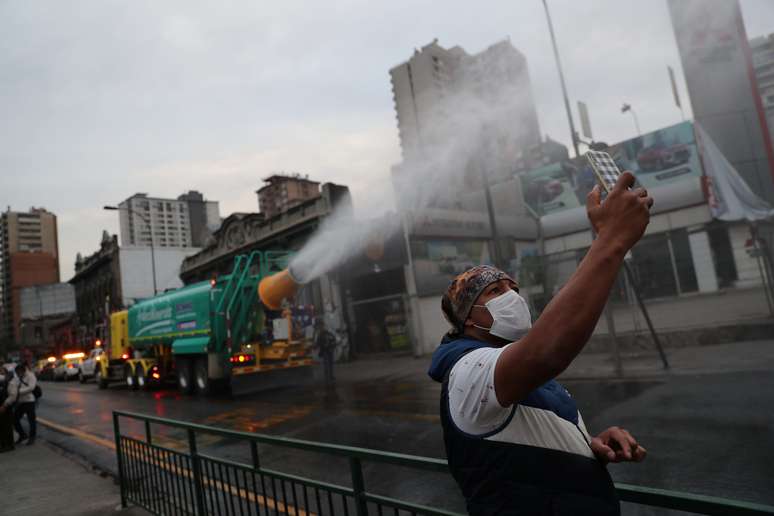 Homem usa telefone celular enquanto um caminhão pulveriza um composto de amônia e água  para desinfetar ruas em meio à pandemia do coronavírus, em Santiago. 12/5/2020. REUTERS/Ivan Alvarado