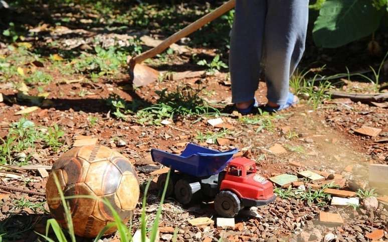 Criança trabalhando com uma enxada ao fundo, na frente uma bola e um carrinho de brinquedo