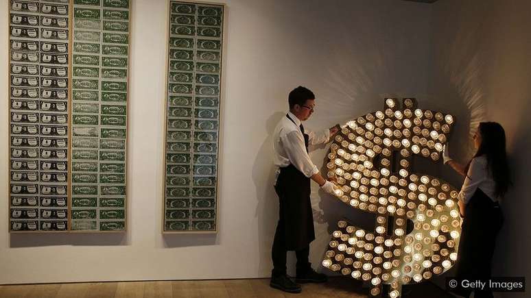 S&iacute;mbolo do d&oacute;lar feito de l&acirc;mpadas e refletores, cria&ccedil;&atilde;o dos artistas Tim Noble e Sue Webster, ao lado de quadros de Andy Warhol