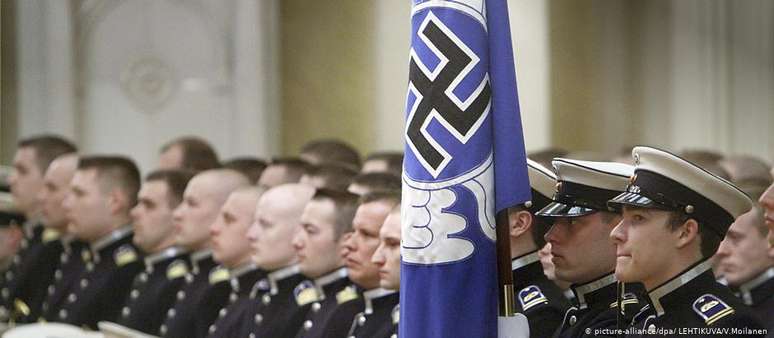 Militares finlandeses ao lado de uma bandeira com uma suástica azul, símbolo ligado à fundação da Força Aérea do país 