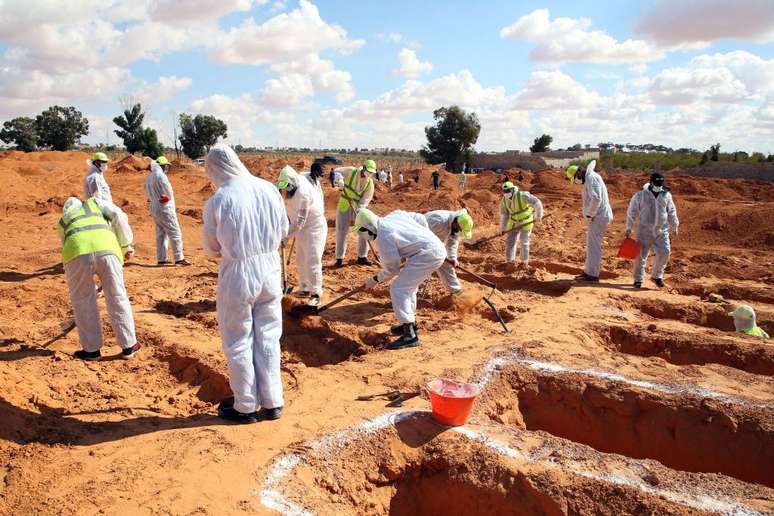Homens em trajes de proteção brancos desenterrando corpos de mortos