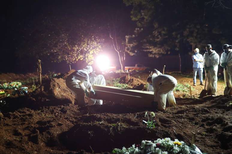 Coveiros vestindo roupas de proteção carregam o caixão de um homem de 32 anos que morreu de doença coronavírus (COVID-19) enquanto holofotes iluminam os túmulos durante os enterros noturnos no cemitério da Vila Formosa em São Paulo