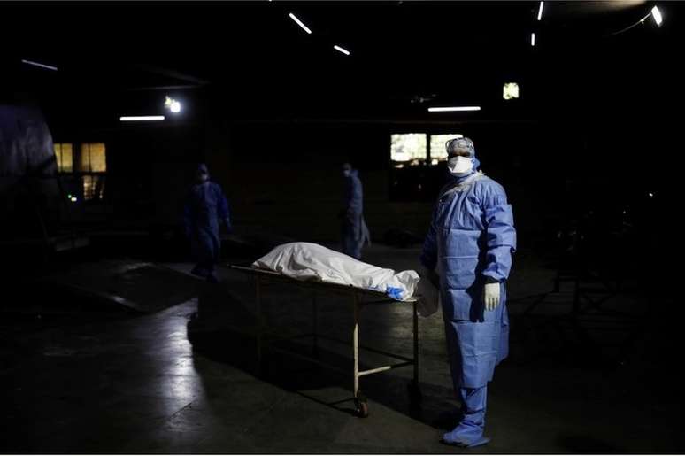 Espera para cremação em Delhi: profissional de saúde ao lado do corpo de um homem que morreu de covid-19