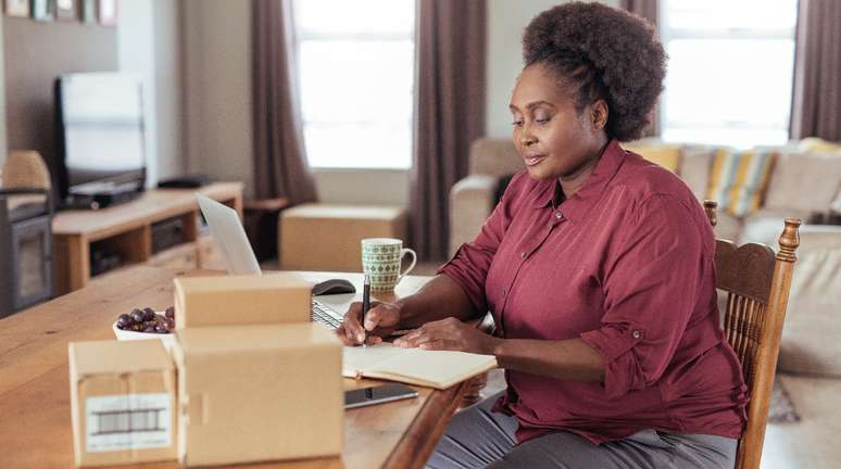 Mulher sentada em cadeira, com um caderno apoiado na mesa enquanto escreve