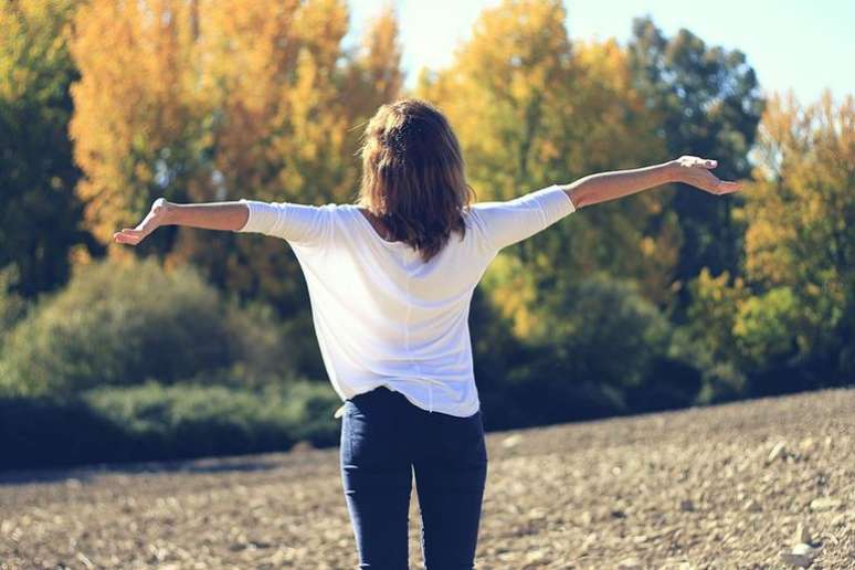 Mulher de braços abertos em campo feliz por sua limpeza astral - Reprodução/Canva.com