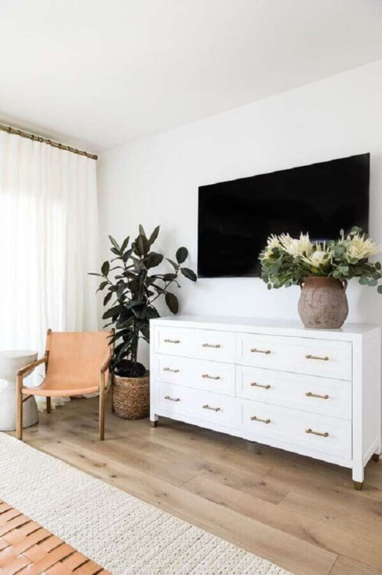 19. Cômoda branca grande para quarto amplo decorado com poltrona de madeira e vaso de planta no chão – Foto: Apartment Therapy