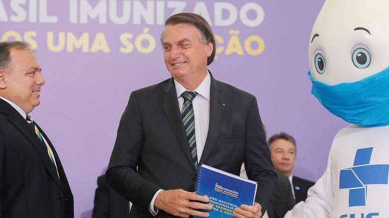 Bolsonaro (ao centro) recebe do ministro da Saúde, Eduardo Pazuello (à esquerda), e do personagem Zé Gotinha (à direita) o Plano Nacional de Operacionalização da Vacinação contra a Covid-19 em evento no dia 16 de dezembro de 2020