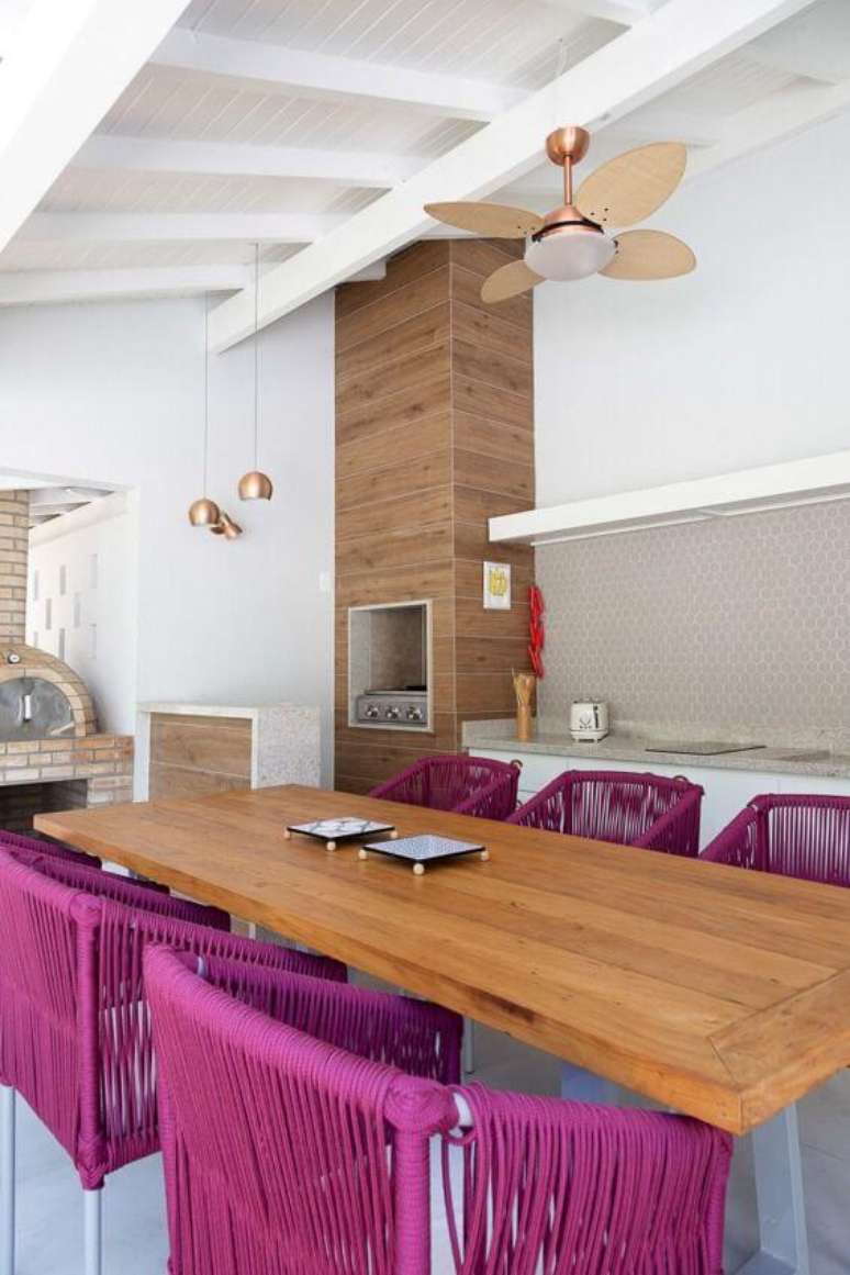 4. Área de lazer moderna com mesa de madeira e cadeiras cor de rosa e churrasqueira pequena no canto – Foto Agata Cunha