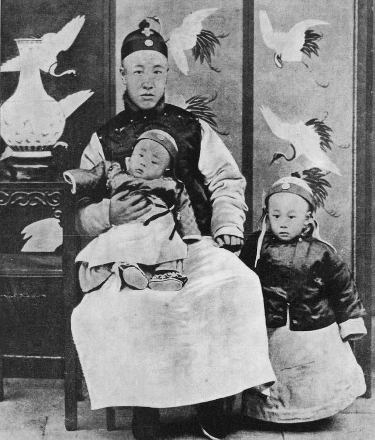1909: Príncipe Chun, regente e controlador da nação, com seu filho mais novo ajoelhado e Puyi (1906-1967), Imperador da China, ao seu lado