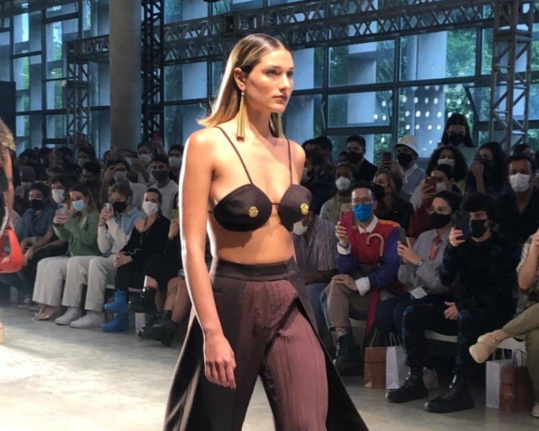 Sasha abriu o desfile com um top tipo biquíni, com detalhe dourado, calça com sobressaia por cima, brincos longos e sandália azul 