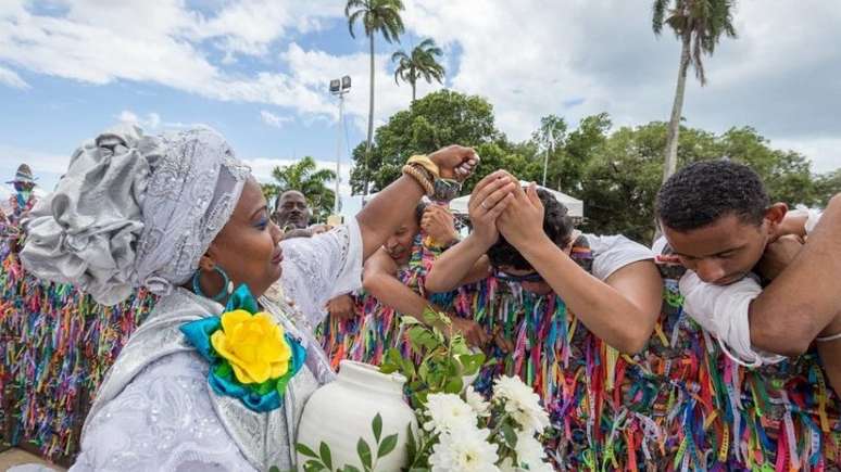 A imagem enquadra cinco pessoas. Em destaque, uma baiana com roupas e turbante cinza. Ela carrega um vaso branco com flores brancas e folhas. A frente dela tem um homem branca com o rosto abaixado e as mãos sob a cabeça, recebendo benção. Ao lado dele, um homem negro também está com a cabeça baixa. Eles estão encostados em um gradil repleto de fitas coloridas do Senhor do Bonfim