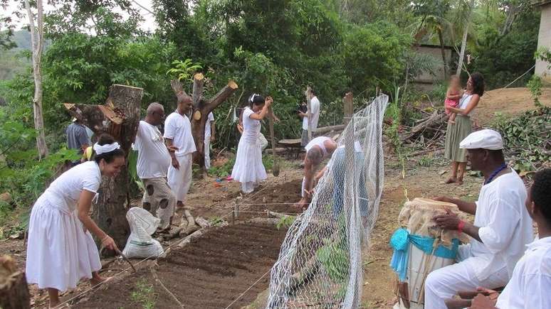 A imagem enquadra pessoas vestidas de branco em um terreno com árvores. No canto esquerdo, cinco pessoas estão preparando uma terra para plantio. No canto direito, à frente, dois homens negros tocam atabaque. Ao fundo, uma mulher carrega uma criança