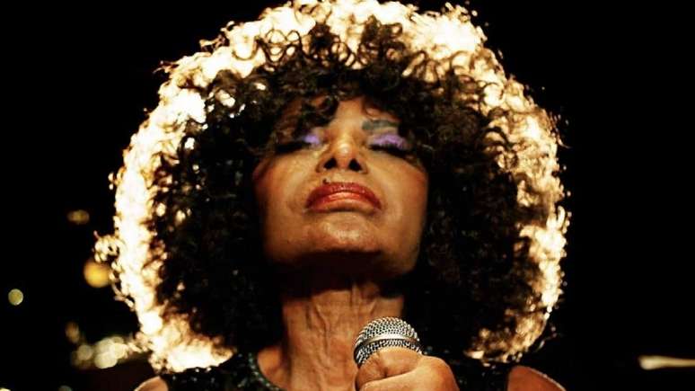 A imagem mostra Elza Soares. A cantora está segurando um microfone, com os olhos fechados e olha para cima