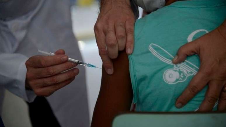Imagem ilustra matéria sobrte vacinação infantil. Uma criança negra é imunizada.