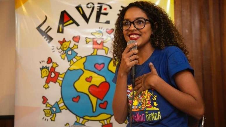 Imagem de divulgação do projeto Favela Mundo. Uma mulher negra e com cabelos cacheados fala ao microfone. Ela veste a camisa do projeto.