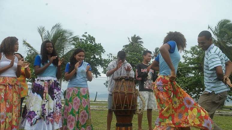 Quatro mulheres quilombolas estão com saias estampadas e três homens batem palmas e tocam um tambor