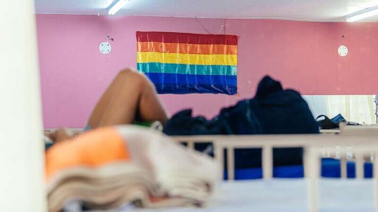 Imagem mostra bandeira LGBTI+ em destaque pendurada em uma parede rosa dentro de um dormitório de uma casa de acolhimento