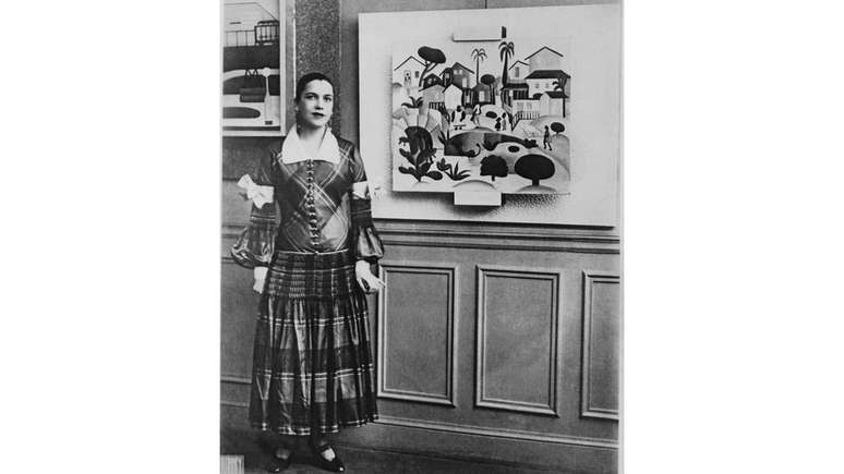 Tarsila do Amaral posa com vestido xadrez em frente ao seu quadro 'Morro de Favela' em vernissage na Galeria Percier, em Paris, em 1926