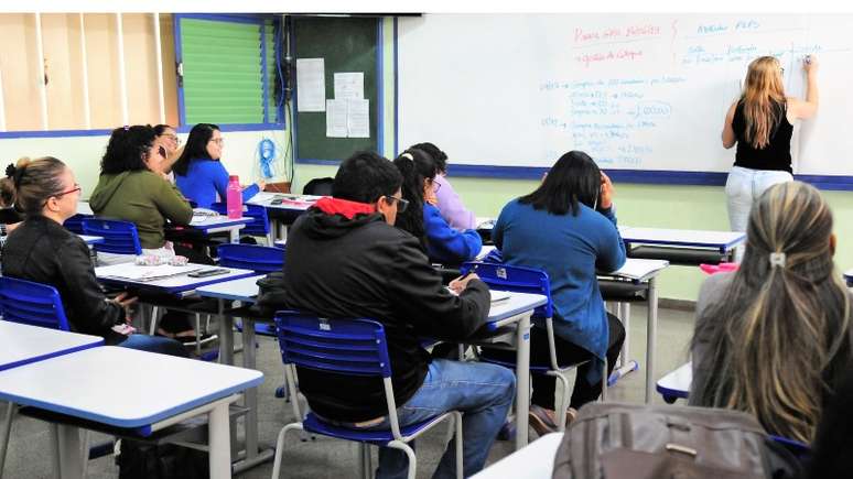 A imagem mostra uma turma de educação para jovens e adultos no Distrito Federal. A professora está de costas escrevendo no quadro branco enquanto os estudantes observam a aula