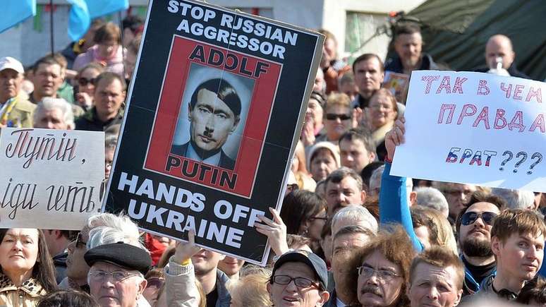 Manifestantes seguram cartaz que retrata o presidente russo, Vladimir Putin, como Adolf Hitler, no qual se lê: 'Detenham o agressor russo, tire as mãos da Ucrânia' — em março de 2014, em Kiev