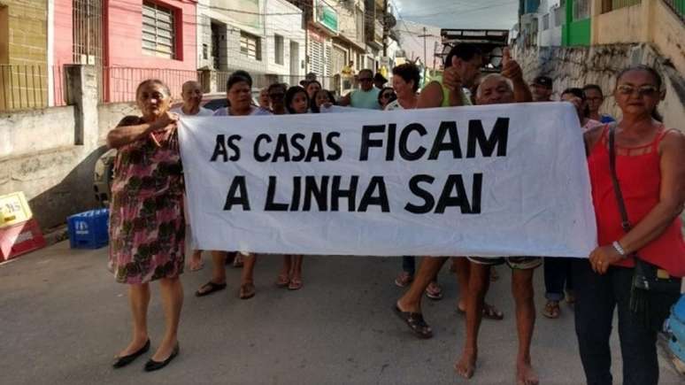 Imagem mostra mulheres negras em ato segurando uma faixa branca com letreiro preto com a frase "a linha sai, as casas ficam"