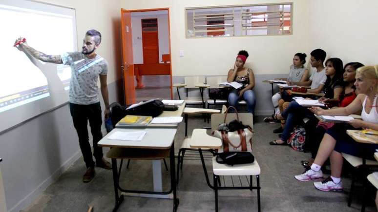 A imagem mostra uma sala de aula com alunos adultos. O professor está a esquerda escrevendo no quadro e os estudantes à direita observam com atenção