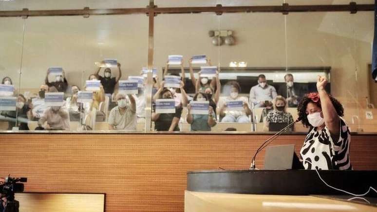 Imagem mostra a vereadora Dani Portela no canto direito falando durante plenária. Ao fundo, cidadãos sentados seguram placas escritas "Rua Marielle Franco"