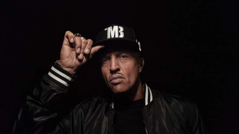 Imagem mostra o rapper Mano Brown em fundo preto segurando um boné da mesma cor e olhando diretamente para a câmera