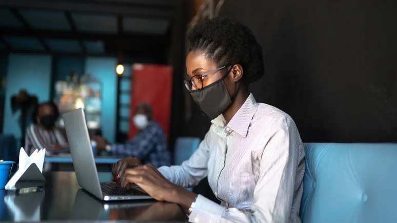 Mulher negra usa uma máscara cirúrgica preta e veste uma camisa social branca enquanto digita algo no notebook.