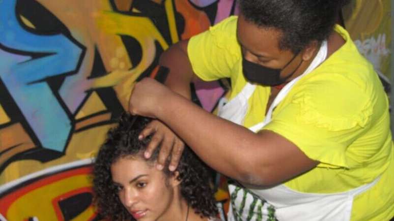 Imagem mostra Amanda NegraSim de amarelo, no canto direito, trançando o cabelo de uma jovem