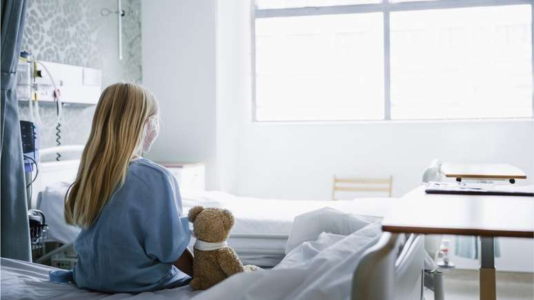 Jovem sentada em uma cama de hospital com um ursinho de pelúcia ao lado dela