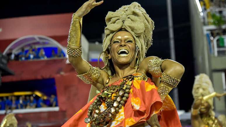 Imagem mostra passista da ala em referência à África, levantando um dos braços e sorrindo. Ela está com adornos étnicos e com maquiagem dourada