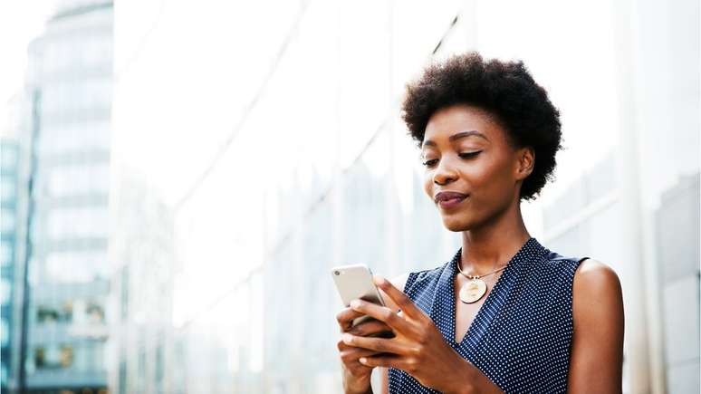 Mulher negra mexe no celular em frente a edifício comercial