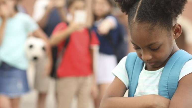 Uma menina negra está cabisbaixa e de braços cruzados enquanto um grupo de crianças ri dela