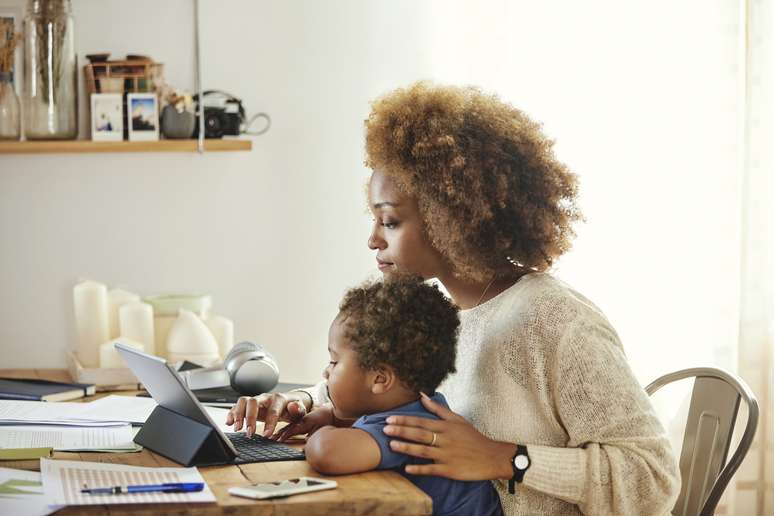 Boy looking at mother using digital tablet. Woman sitting with son at table in kitchen. She is working from home.