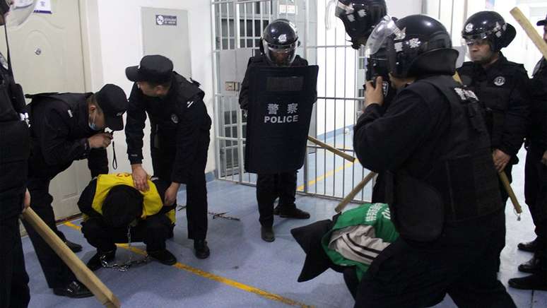 Um homem detido aparece agachado, de cabeça baixa, rodeado por policiais com cacetetes