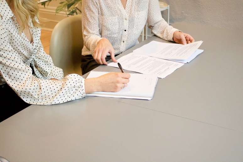 Foto mostra duas mulheres sentadas &agrave; mesa com pap&eacute;is e uma delas escrevendo no papel, indicando uma assinatura de fechamento de neg&oacute;cio.
