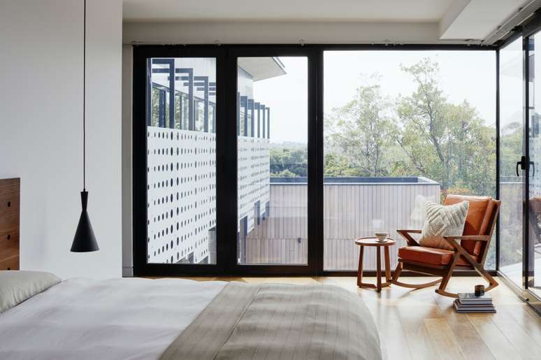 Foto mostra um quarto de casal com uma janela de vidro que toma toda a parede lateral da cama. H&aacute; uma poltrona de madeira e estofado laranja, com mesinha lateral perto da janela.
