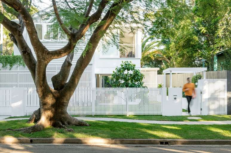 Foto mostra fachada frontal da casa, toda branca, com muito verde e homem saindo pelo port&atilde;o de entrada. Em primeiro plano, h&aacute; uma grande &aacute;rvore na cal&ccedil;ada.