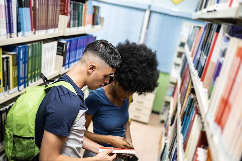 Dois rapazes estão folheando um livro em um corredor de uma biblioteca. Um é branco de cabelo curto e outro é negro, com cabelo médio