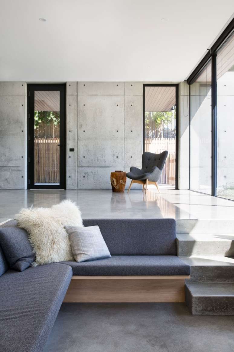 Sala de estar rebaixada com sof&aacute;s na cor azul, ch&atilde;o e paredes cimentadas. Poltrona cinza ao fundo.