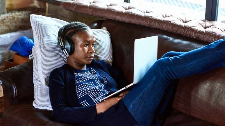 Mulher usando laptop e fones de ouvido enquanto est&aacute; deitada em um sof&aacute;