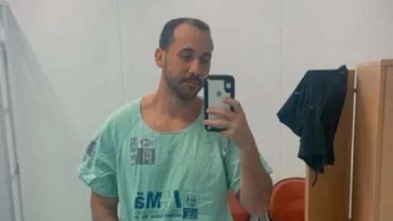 Giovanni está na frente de um espelho. Ele segura um celular e usa um uniforme de hospital