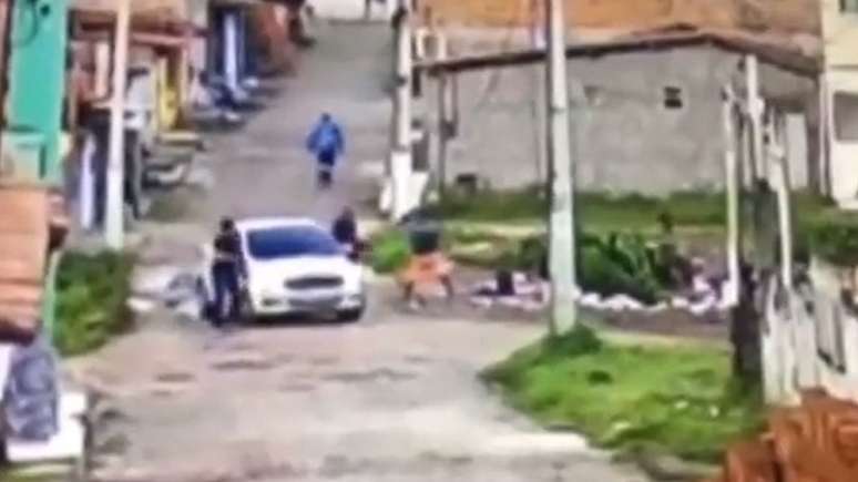 Imagem mostra dois policiais ao lado de um carro e um homem com as mãos na cabeça