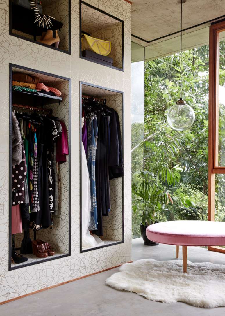 Closet com nichos para roupas e acessórios, um tapete branco felpudo e um banco com design orgânico na cor rosa claro. A parede de vidro dá vista para a floresta.
