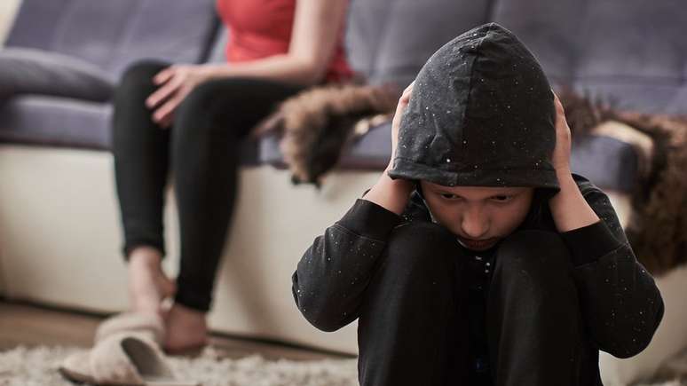 Crian&ccedil;a encapuzada cobre os ouvidos e no fundo h&aacute; uma mulher adulta sentada em um sof&aacute;
