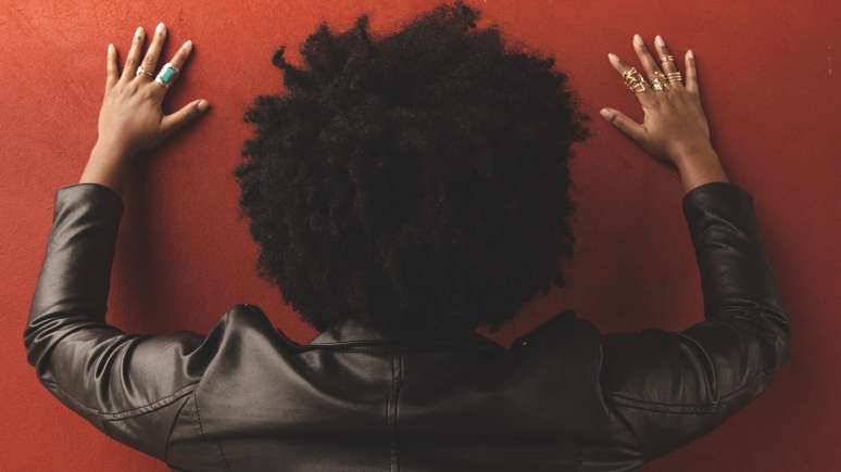 Imagem mostra uma mulher negra de costas, com as mãos na parede como em um enquadro policial.