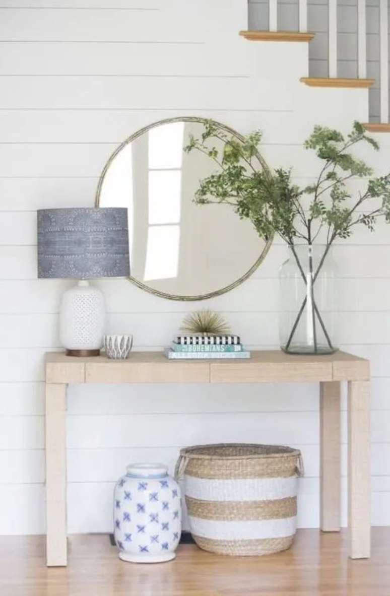 Hall de entrada com mesa de console de madeira. Sobre ela jaz um vaso com uma grande planta e um abajur num arranjo assimétrico. Na parede está um espelho circular.